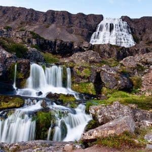 Ein mehrstufiger Wasserfall stürzt einen felsigen Berg hinab, wobei kleinere Wasserfälle im Vordergrund über moosbewachsene Felsen fließen.