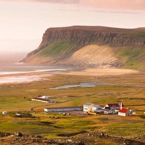Ein kleines Küstendorf mit einer Kirche liegt auf einer grünen Ebene zwischen einem breiten Strand und einer großen, steilen Klippe.