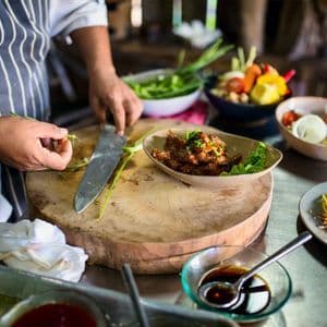 Ein Koch in einer gestreiften Schürze bereitet Zutaten auf einem runden Holzschneidebrett in einer Küche voller Schüsseln mit Lebensmitteln zu.