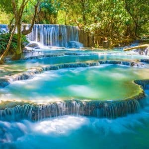 Ein breiter, mehrstufiger Wasserfall fließt in mehrere türkisfarbene Becken in einem sonnendurchfluteten grünen Wald.