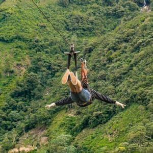 Eine Person seilrutscht kopfüber mit ausgestreckten Armen über ein üppiges, grünes, bewaldetes Tal.