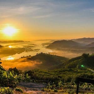 Goldener Sonnenaufgang über einem breiten Fluss, der sich durch neblige, dschungelbedeckte Berge schlängelt, gesehen von einem hölzernen Aussichtspunkt im Vordergrund.