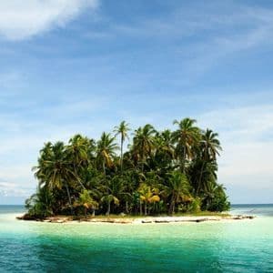 Una pequeña isla tropical cubierta de palmeras, rodeada de aguas turquesas y cristalinas bajo un cielo azul con nubes ligeras.
