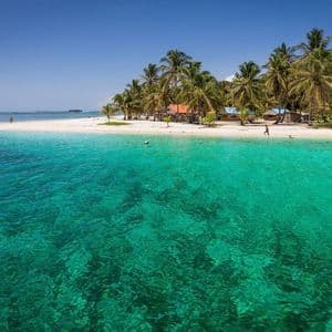 Klares türkises Wasser im Vordergrund mit einem weißen Sandstrand und Palmen auf einer Insel unter blauem Himmel.