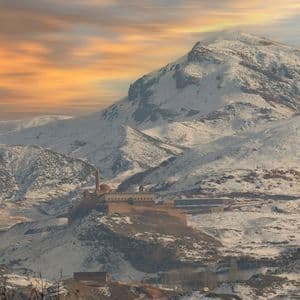 Un palazzo storico e un minareto si ergono su una collina circondata da vaste montagne innevate sotto un cielo colorato al tramonto.