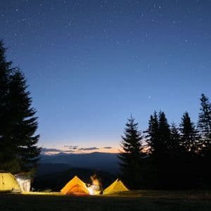 Un campeggio con tre tende illuminate e un falò in una radura nel bosco sotto un cielo notturno stellato.