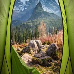 Una vista in prima persona dall'interno di una tenda verde, che si affaccia su una valle boscosa e montagne frastagliate e innevate.
