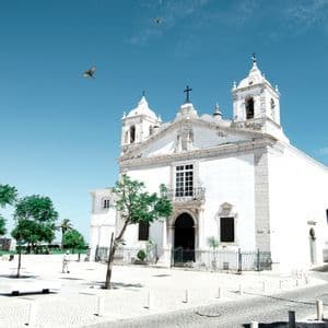 A white church with two bell towers stands in a cobblestone plaza under a clear blue sky with birds flying overhead.