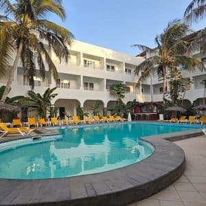 A turquoise swimming pool surrounded by yellow lounge chairs and palm trees in the courtyard of a white hotel building.