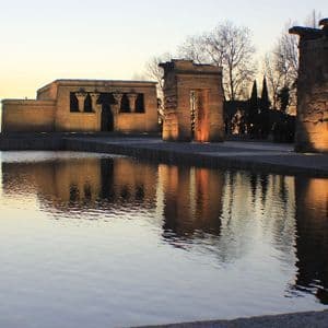 An ancient stone temple is illuminated and reflected in a long pool of water at sunset.