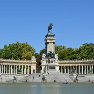 A large monument with an equestrian statue and a colonnade overlooks a lake with people rowing boats under a clear blue sky.