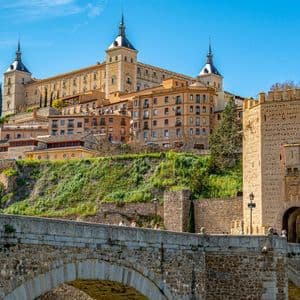 A large stone fortress sits on a green hill above a historic arched bridge and fortified gate under a clear blue sky.