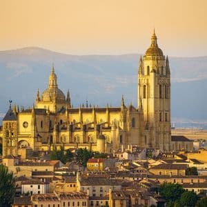 A grand Gothic cathedral with domes and a bell tower sits above a town at sunrise, with hazy mountains in the background.