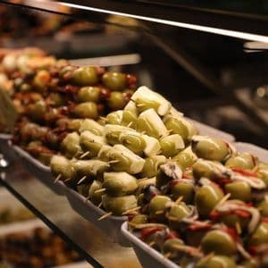 Trays of tapas skewers with green olives, cheese, and anchovies sit on a glass shelf in a display case.