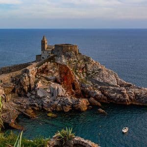 Una chiesa di pietra con un campanile sorge su una penisola rocciosa che si protende nel mare blu, con una piccola barca sotto.
