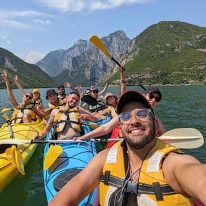 Un gruppo WeRoad si scatta un selfie sorridente in kayak su un lago con montagne verdi sullo sfondo.