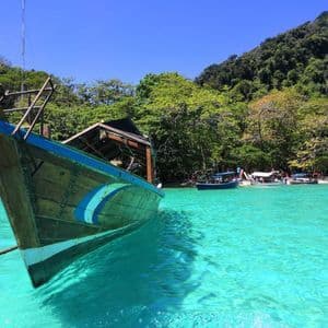 The bow of a traditional wooden boat moored in bright turquoise water, with a lush, forested island in the background under a clear sky.