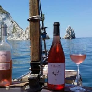 Due bottiglie di vino rosato e due bicchieri sul ponte di una barca a vela in mare, con scogliere sullo sfondo.