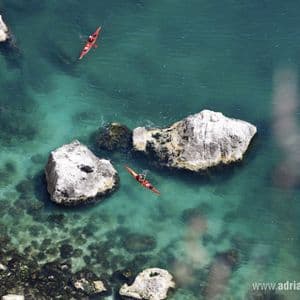Vista aerea di due persone su kayak rossi che remano in acque turchesi e cristalline vicino a una costa rocciosa.