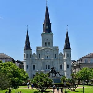 A large white cathedral with three steeples stands behind a green park containing an equestrian statue and people walking.