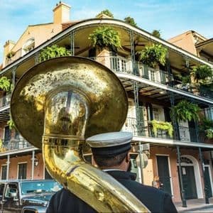 A musician from behind carries a large brass tuba on a street in front of a building with ornate, plant-filled balconies.
