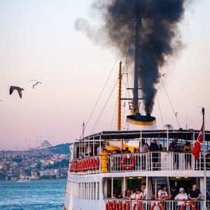 A multi-deck ferry with passengers sails on the water, releasing a thick plume of black smoke from its smokestack against a pale sky.