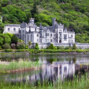 Un gran castillo de piedra se alza a la orilla de un lago tranquilo, con su reflejo visible en el agua y un exuberante bosque verde elevándose detrás.