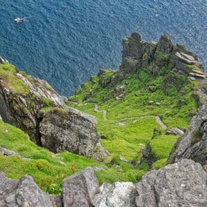 Una vista panorámica desde un acantilado rocoso que domina una empinada ladera verde, con un sendero de piedra serpenteante que desciende hasta el profundo mar azul, donde se divisa una pequeña embarcación.