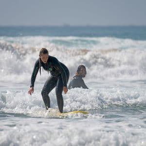 A woman in a wetsuit on a WeRoad group trip surfs on a yellow board, with another person smiling in the background waves.