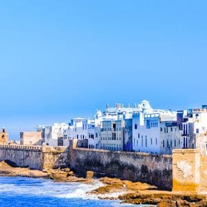 A fortified coastal city with whitewashed buildings stands on a rocky shore, with blue ocean waves in the foreground under a clear sky.