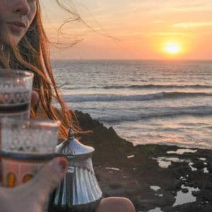 Two people toast with traditional tea glasses on a rocky coast as the sun sets over the ocean.