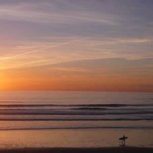 A silhouetted surfer carrying a surfboard walks along the shoreline during an orange sunset over the ocean.