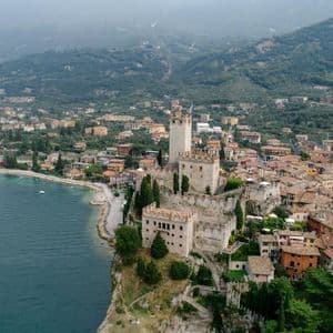 An aerial view of a stone castle on a cliff overlooking a lakeside town, with green mountains in the background.