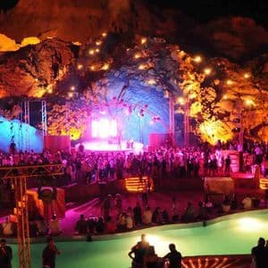 Una grande folla di persone a un evento notturno con piscina e palco, sullo sfondo di formazioni rocciose illuminate drammaticamente.