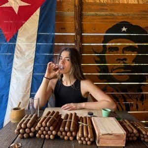 Une femme fume un cigare à une table couverte de paquets de cigares, avec un drapeau cubain et une fresque de Che Guevara en arrière-plan.