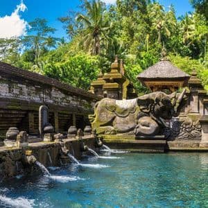 Steinerne Brunnen speien Wasser in einen klaren blauen Pool an einem moosbewachsenen Tempel mit einer großen Elefantenstatue und Dschungelkulisse.