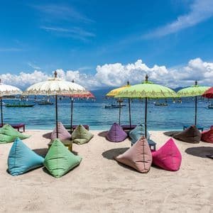 Bunte Sitzsäcke und traditionelle Sonnenschirme säumen einen Sandstrand vor einem türkisfarbenen Meer mit Booten unter blauem Himmel.