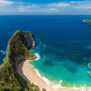 Eine Luftaufnahme einer großen, bewachsenen Felsformation neben einem weißen Sandstrand mit türkisfarbenem Wasser.