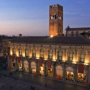 Una vista dall'alto di una piazza storica cittadina al crepuscolo, con edifici illuminati e un'imponente torre in mattoni
