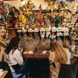 Deux femmes participant à un voyage de groupe WeRoad peignent des masques à une table dans un atelier rempli de masques vénitiens.