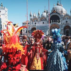 Une foule en costumes colorés et masques à plumes célèbre un carnaval sur une place historique.