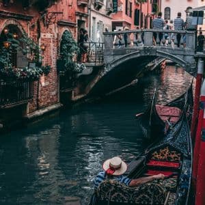 A gondolier in a straw hat steers a gondola down a narrow canal between brick buildings, with a stone bridge in the background.
