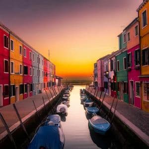 Small boats are moored in a canal between rows of colorful buildings under a glowing sunset sky.