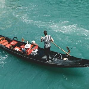 A gondolier in a striped shirt rows a gondola with two passengers from a WeRoad group trip across turquoise water.