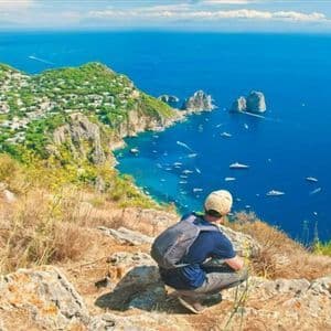 Una persona con mochila se agacha en lo alto de un acantilado rocoso, contemplando un mar azul intenso con barcos, farallones y un pueblo costero.