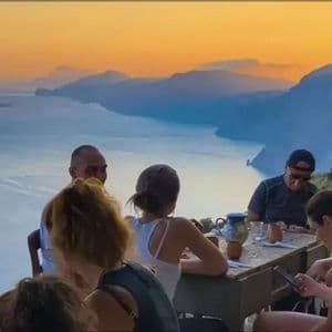 Un viaje en grupo de WeRoad se reúne en una mesa de madera con vistas a una costa montañosa y al mar al atardecer.