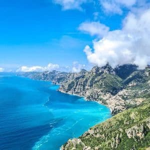 Vista aérea de una costa escarpada y verde que se une a un mar turquesa vibrante bajo un cielo azul con nubes blancas.