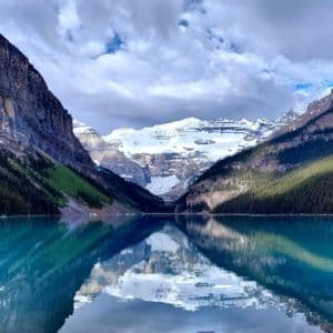 Snow-capped mountains and pine forests are reflected in the calm, turquoise water of an alpine lake under a cloudy sky.