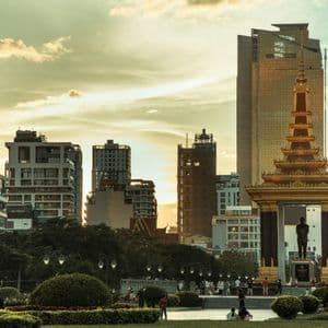 Un monumento dorado con una estatua se alza en un parque contra el horizonte de una ciudad moderna al atardecer.