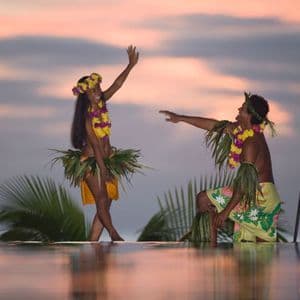 Ein Mann und eine Frau in traditioneller polynesischer Kleidung tanzen bei Sonnenuntergang an einem Infinity-Pool mit einer beleuchteten Tiki-Fackel.